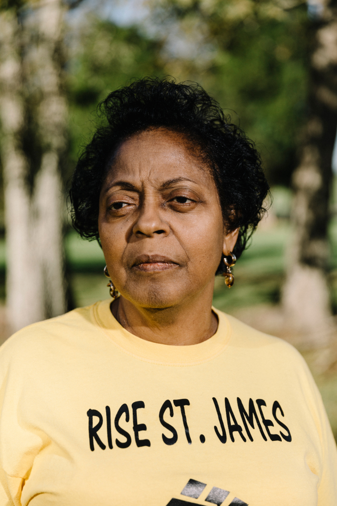 Image description: A portrait of an older Black woman, standing outside. Her hair is tied up, and she wears earrings and a yellow tshirt that reads "RISE St. James."