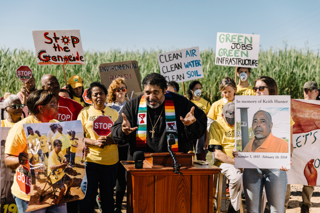 Image description: Photo of a man standing at a podium, speaking into a microphone and gesturing with his hands. Behind him stands a group of people, holding signs that say "Stop the genocide," "Clean air clean water clean soil," and "Green jobs green infrastructure." Most are wearing yellow t shirts that say "RISE St. James."