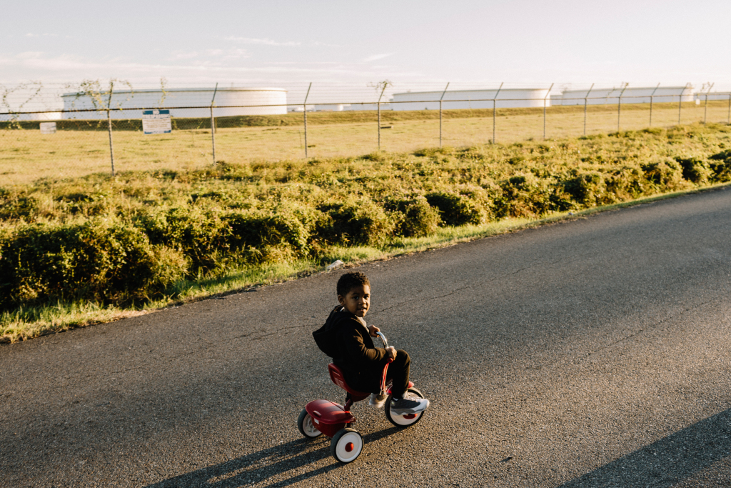 Image description: A young boy riding a tricycle down a road looks at the camera. Behind him is a fenced-in field, and several large factory buildings stand in the distance.